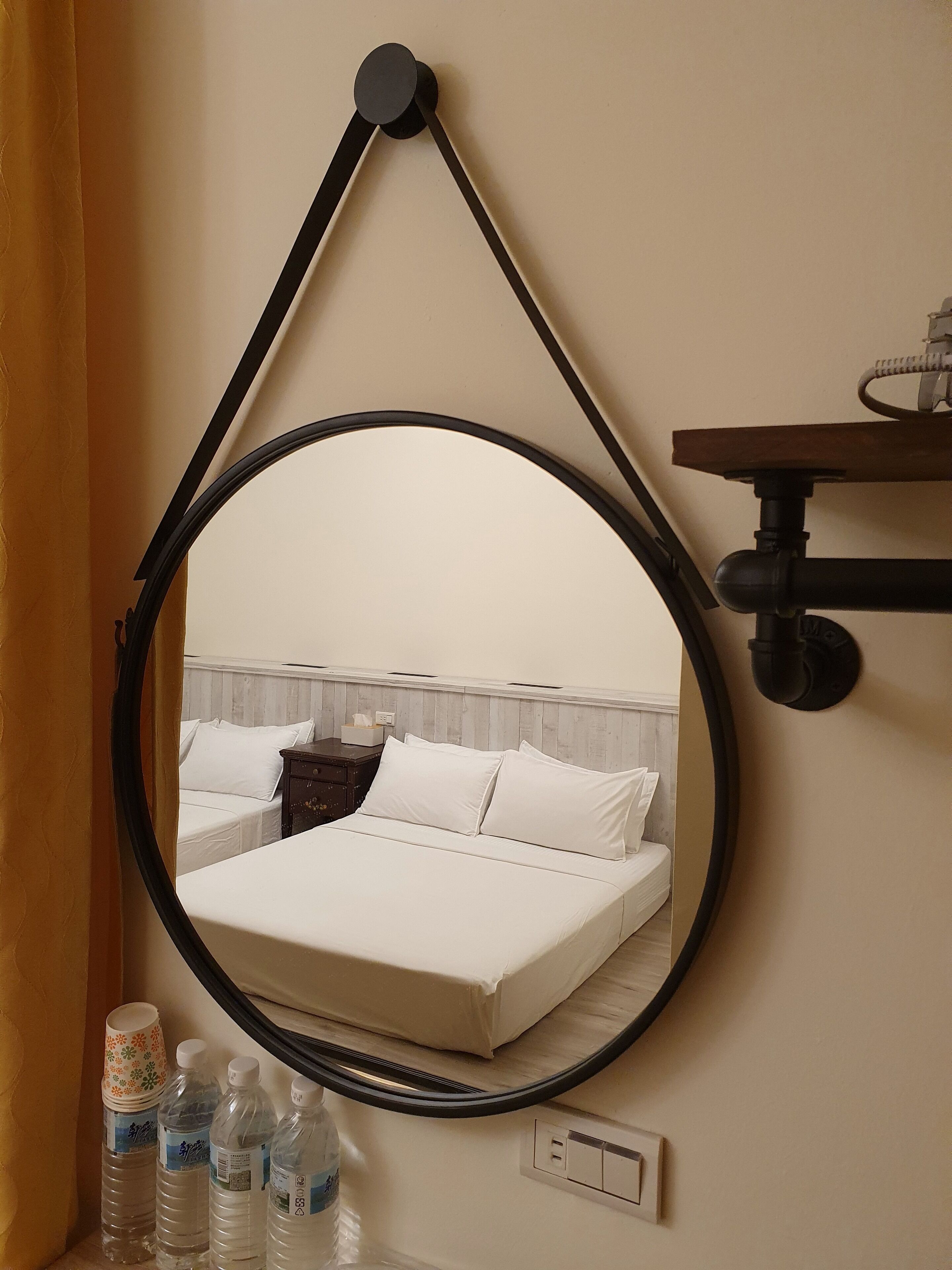 Ein runder Spiegel mit schwarzem Rahmen an der Wand, der ein Bett mit weißer Bettwäsche und ein Holz-Kopfende reflektiert.