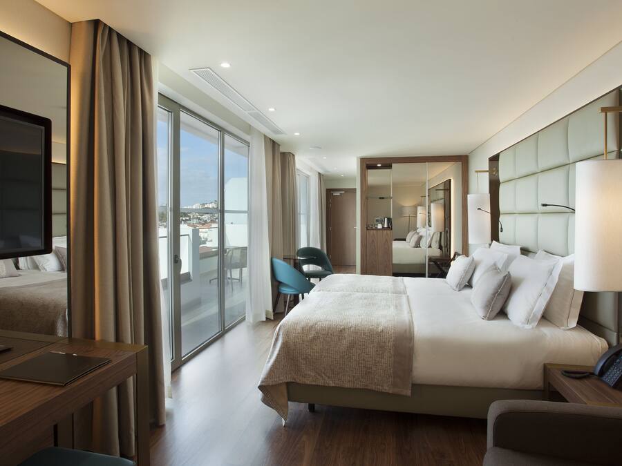 A modern hotel room with a large bed, a desk, a chair, a TV, and a balcony with a view of the city.