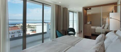 Quarto de hotel com cama grande, uma cadeira, escrivaninha e vista para a cidade e o mar.