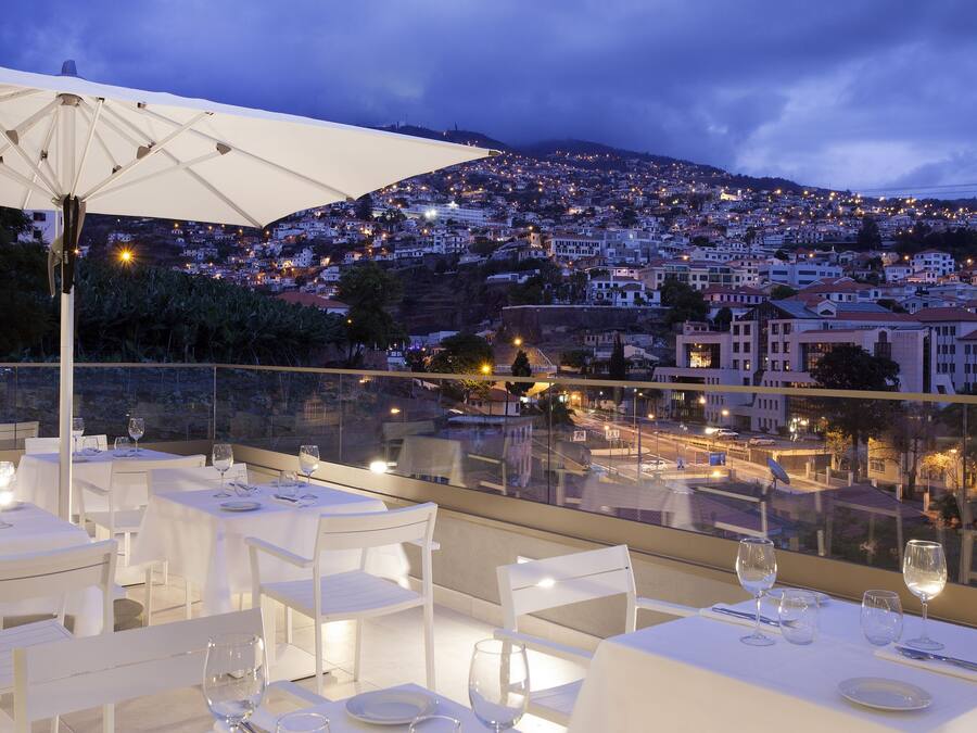 A rooftop dining area with tables set for dinner, overlooking a cityscape at dusk.