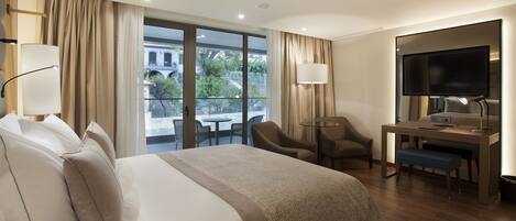 A hotel room with a large bed, a desk with a computer, a TV, and a balcony with a view of buildings and trees.