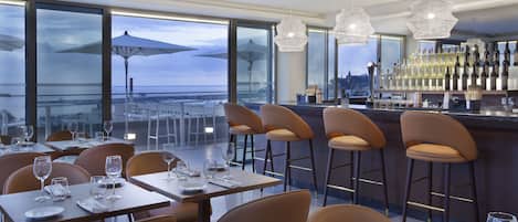 Un restaurante con zona de bar, ventanas amplias con sombrillas y vistas a la ciudad.