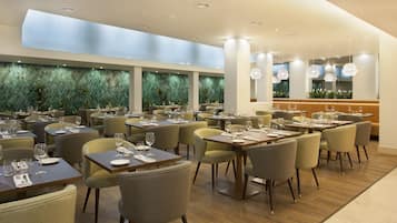 Un restaurant moderne doté de nombreuses tables et chaises, d’un plafond haut avec des luminaires encastrés et d’un mur recouvert de carreaux verts et agrémenté de plantes.