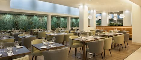 Un restaurant moderne doté de nombreuses tables et chaises, d’un plafond haut avec des luminaires encastrés et d’un mur recouvert de carreaux verts et agrémenté de plantes.