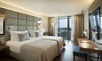 A hotel room with two beds, a desk, a chair, and a large window offering a city view.