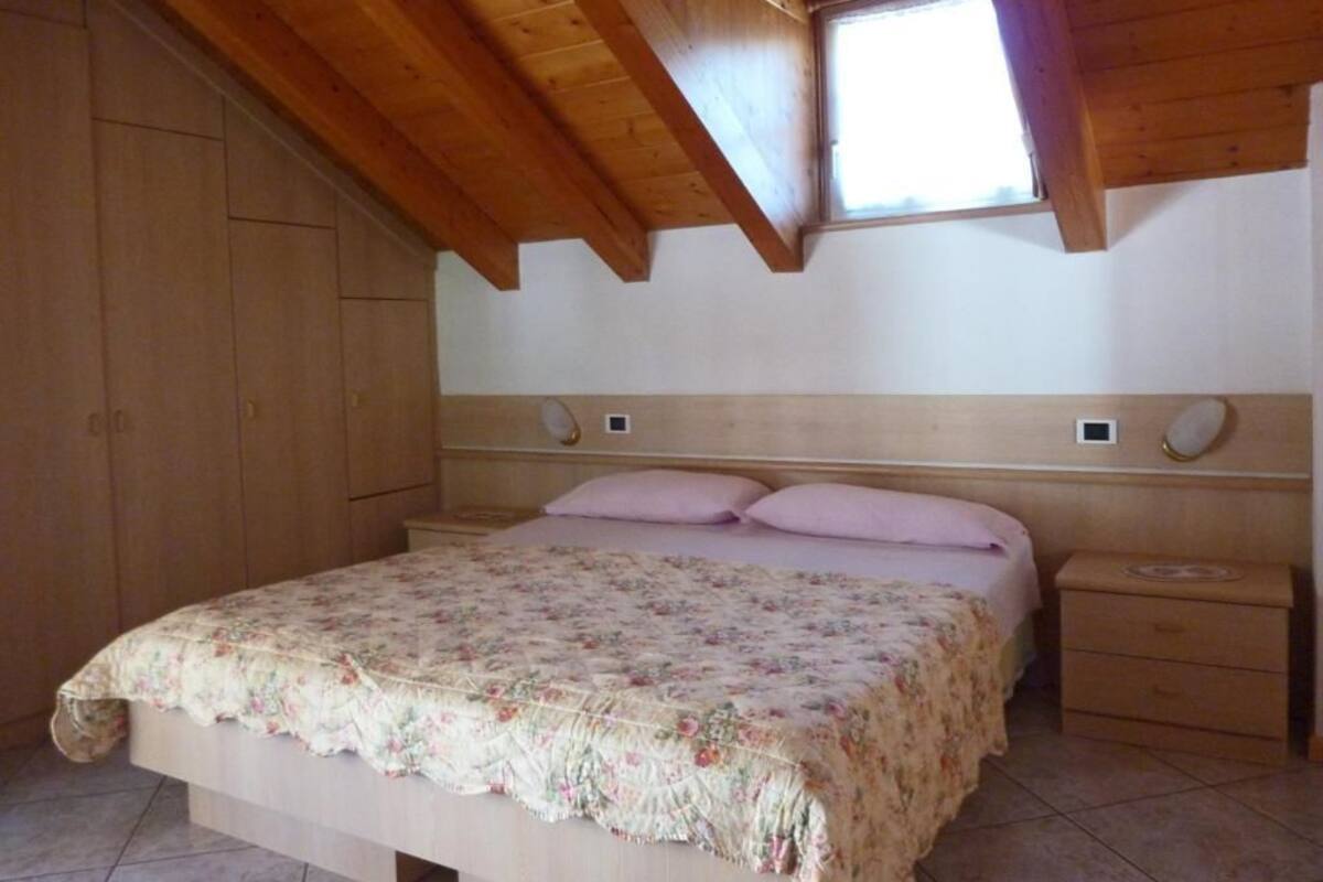 Een slaapkamer met een houten plafond, een raam, een bed met bloemenbedekking, een nachtkastje en een houten garderobe.