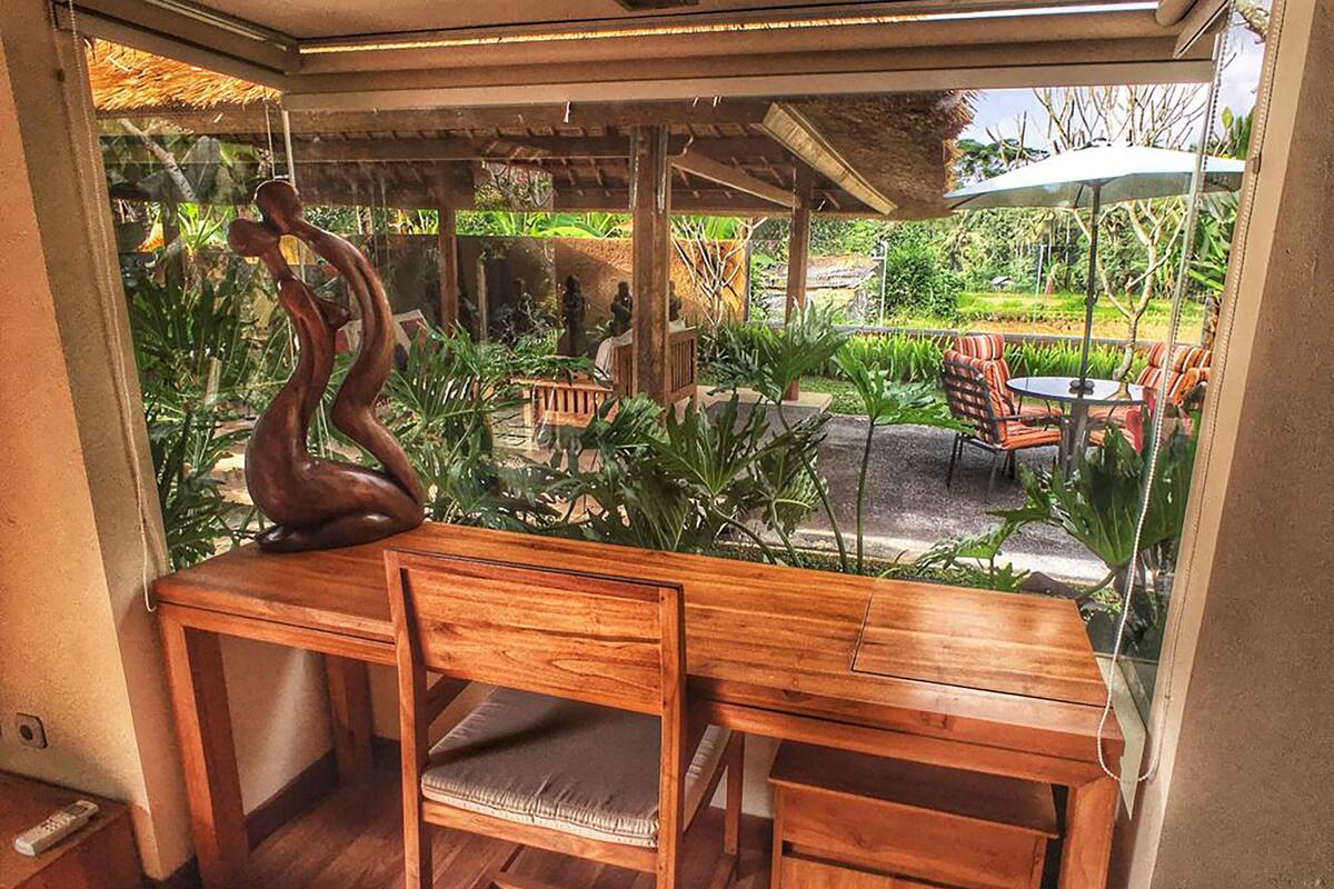 A wooden desk with a chair in a room with a glass wall overlooking a garden with outdoor furniture.