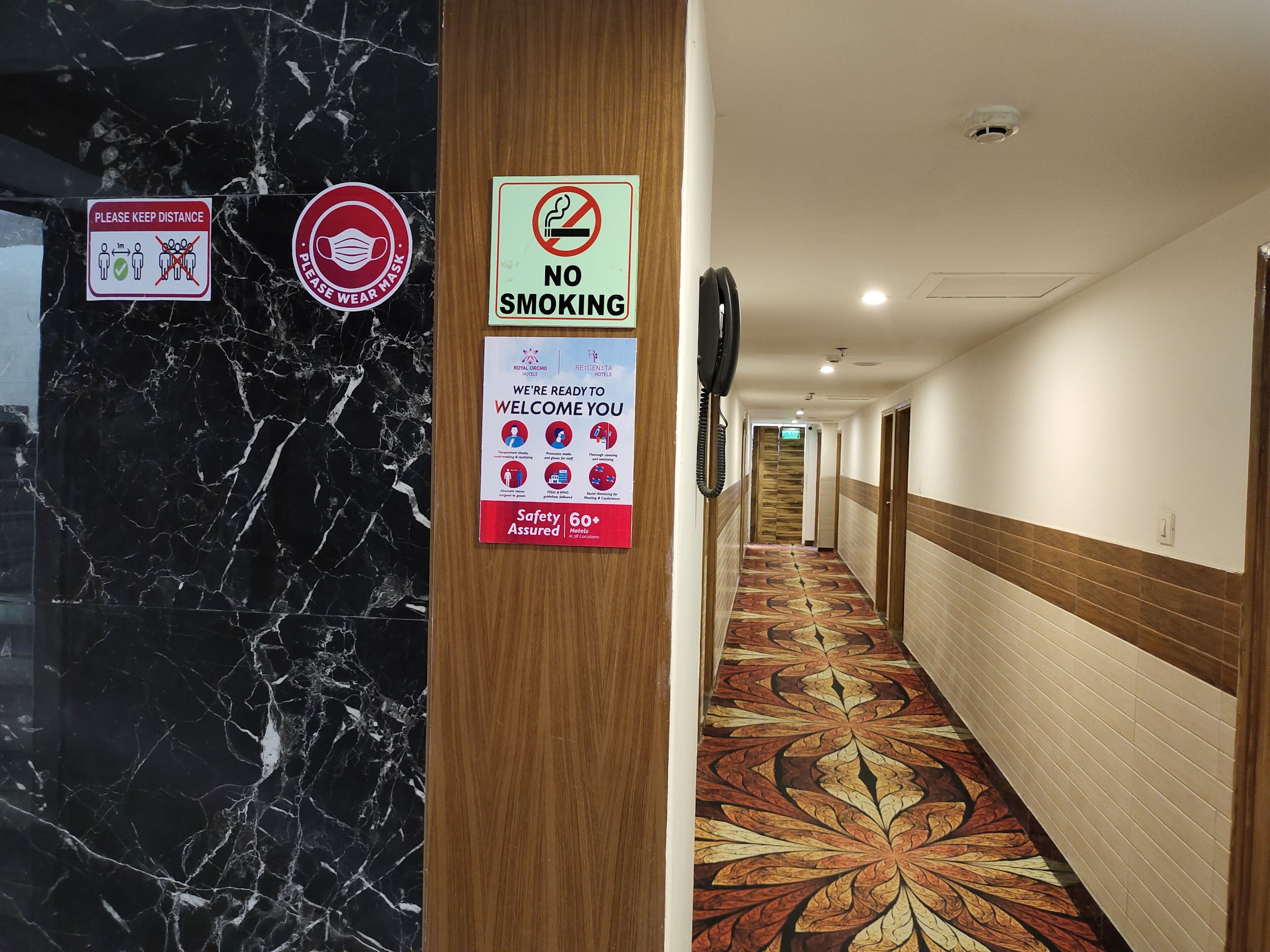 Un pasillo de hotel con paredes de mármol, un cartel de prohibido fumar y un letrero de bienvenida con información sobre seguridad.