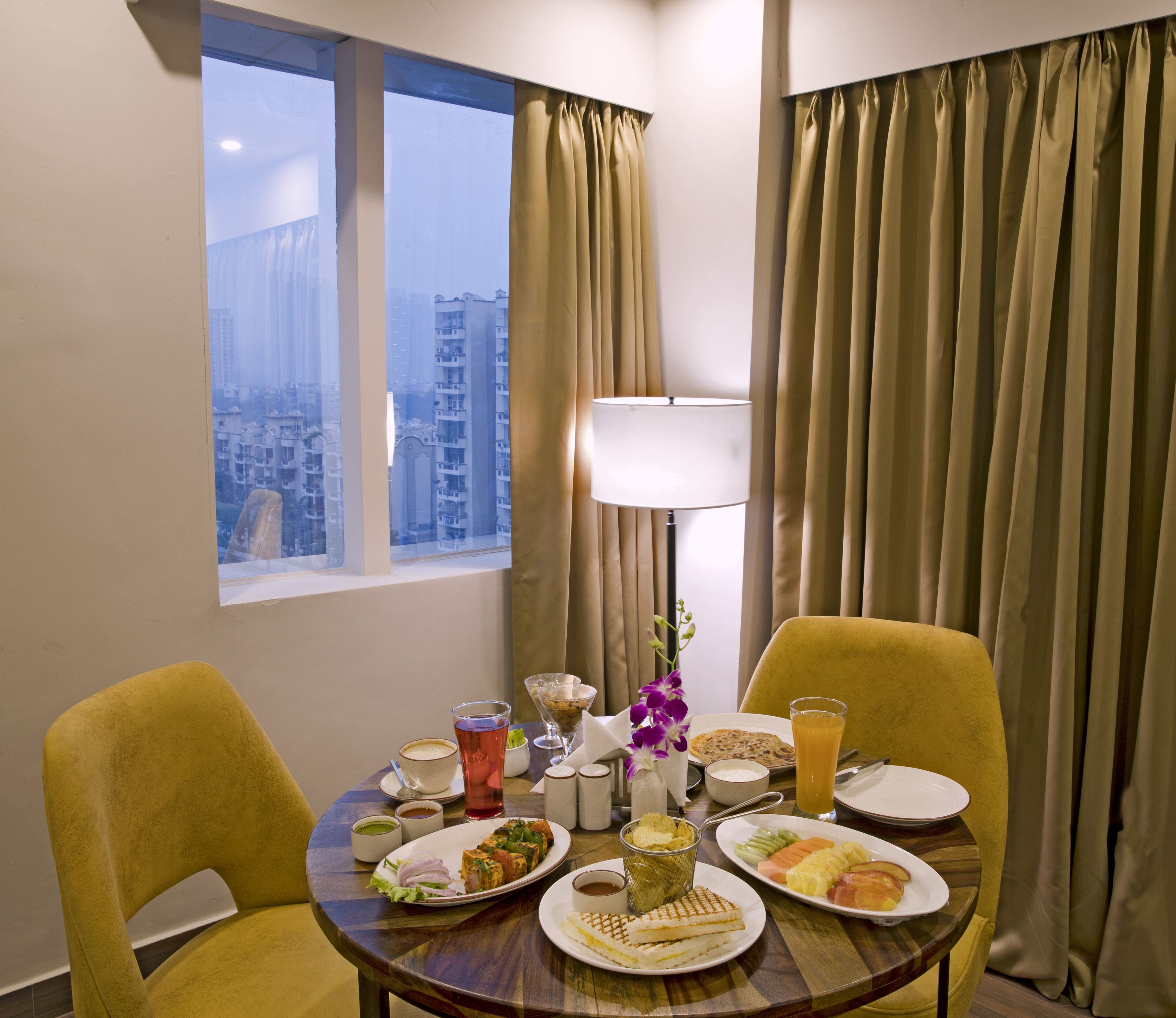 A hotel room with a dining table set for breakfast, featuring plates of food, a lamp, and a view of the city through the window.
