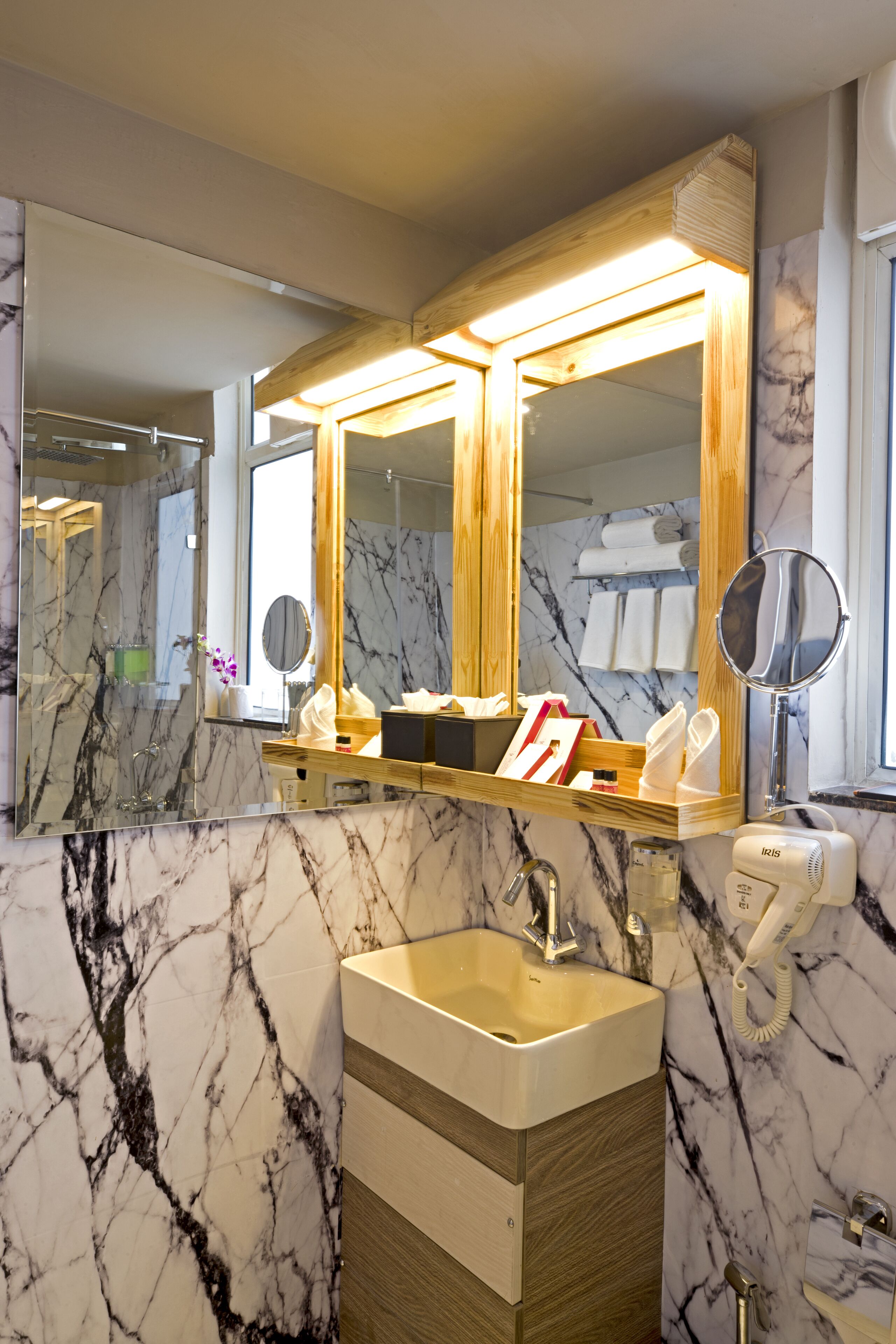 A bathroom with marble walls, a wooden vanity, a sink, and a mirror with lights.