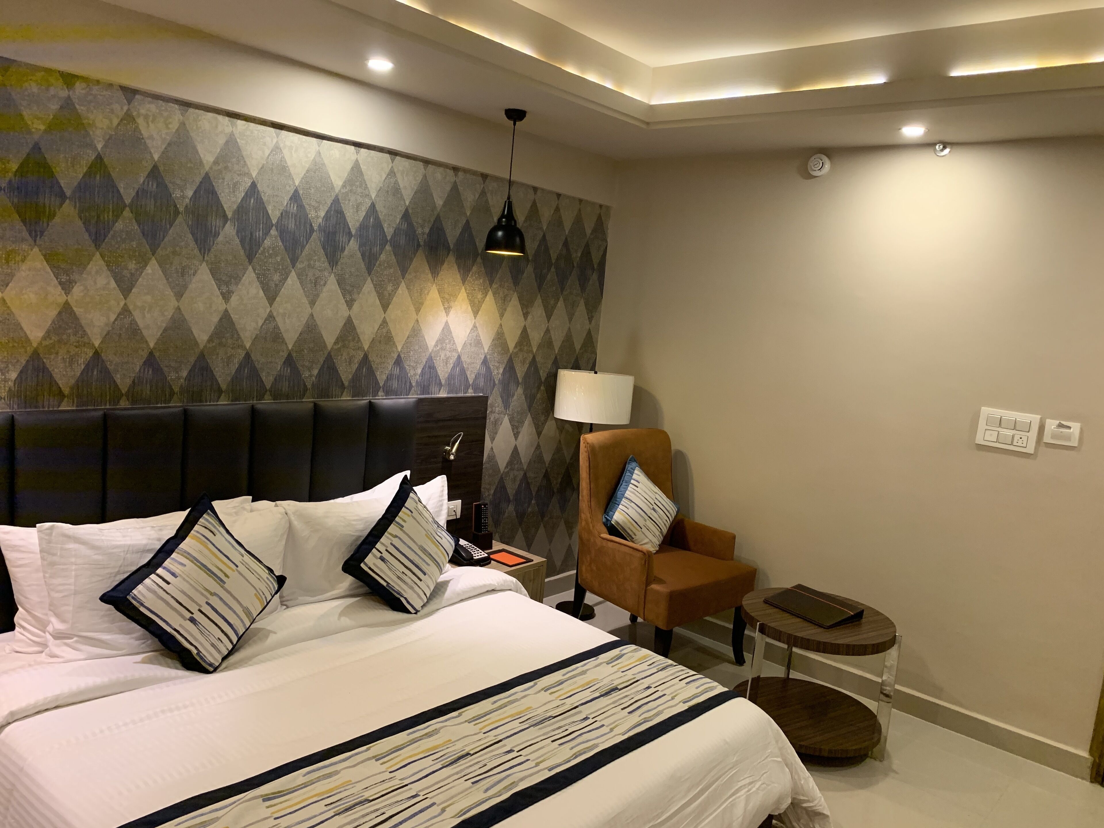 A hotel room with a large bed, a headboard, a nightstand, a lamp, a chair, and a small table.