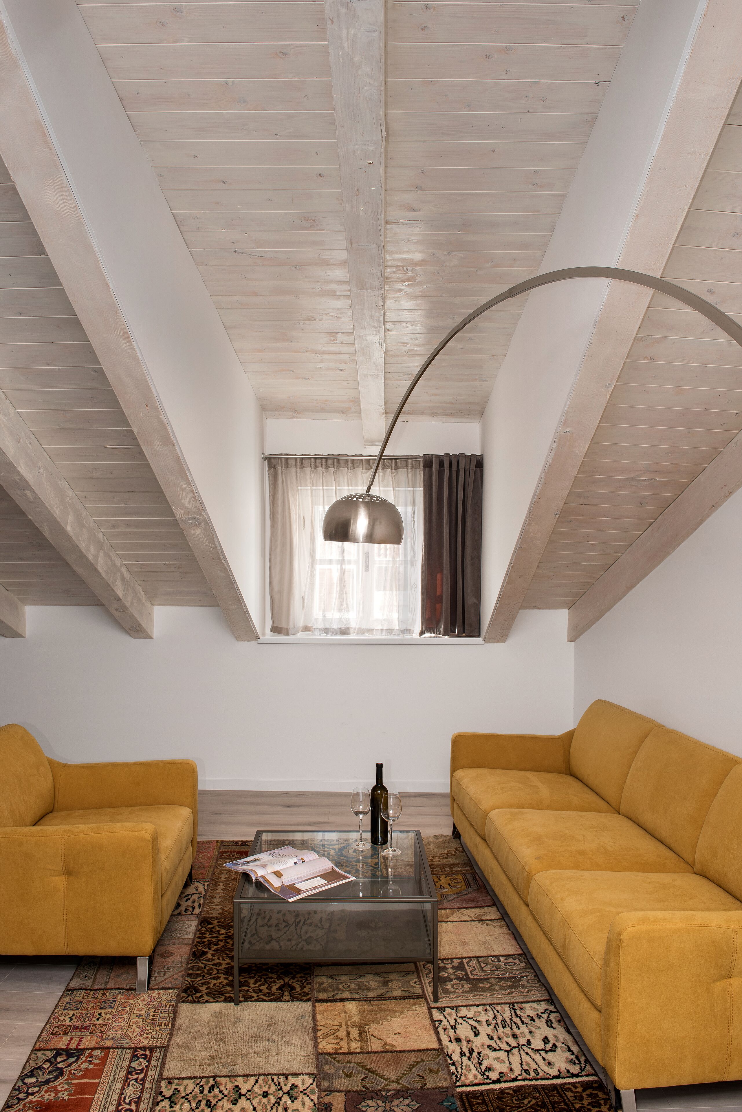 Una stanza con soffitto spiovente, due divani gialli, un tavolino da caffè in vetro e un tappeto decorato.