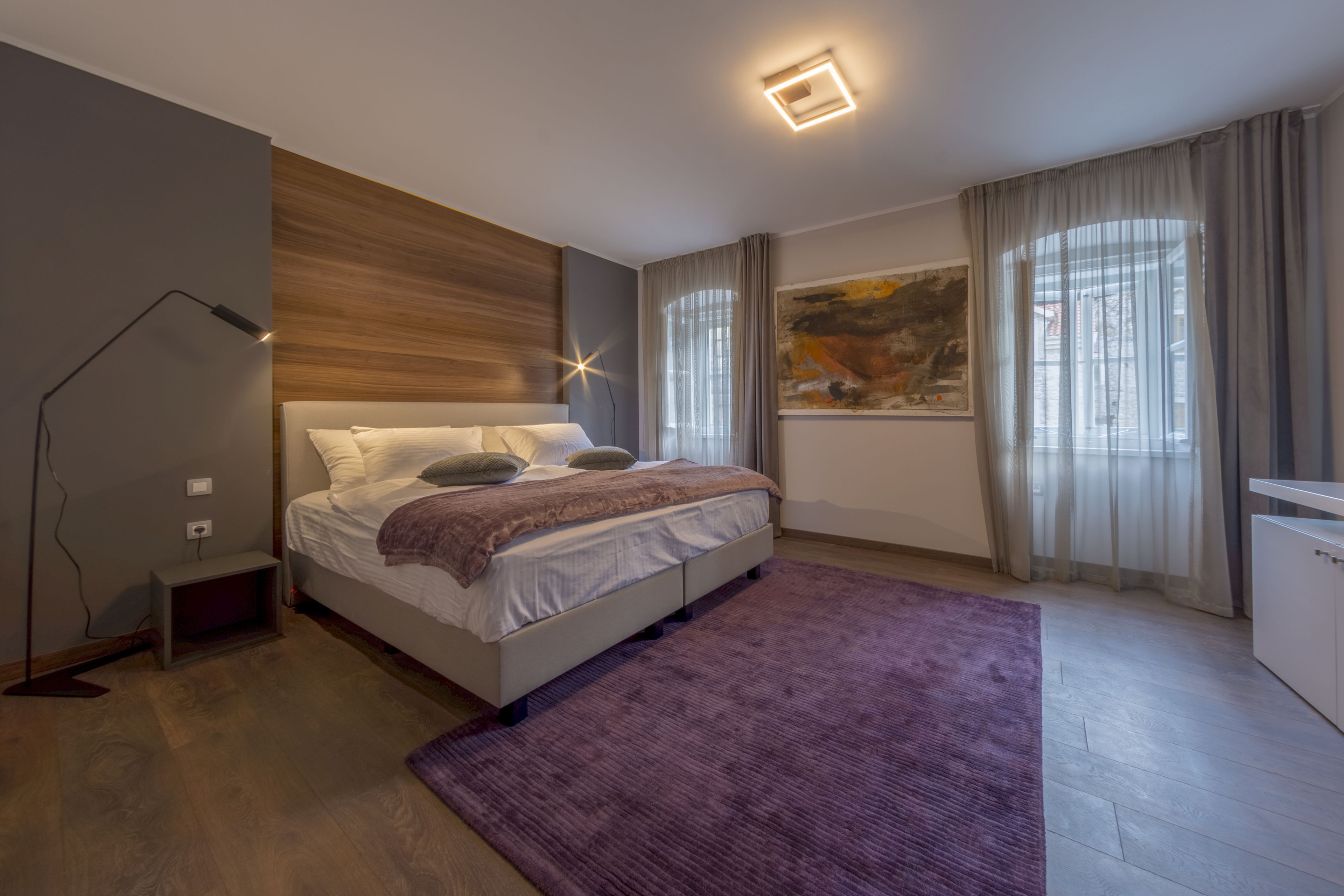 Un dormitorio moderno con una cama, una lámpara de noche, una ventana con cortinas y un cuadro en la pared.