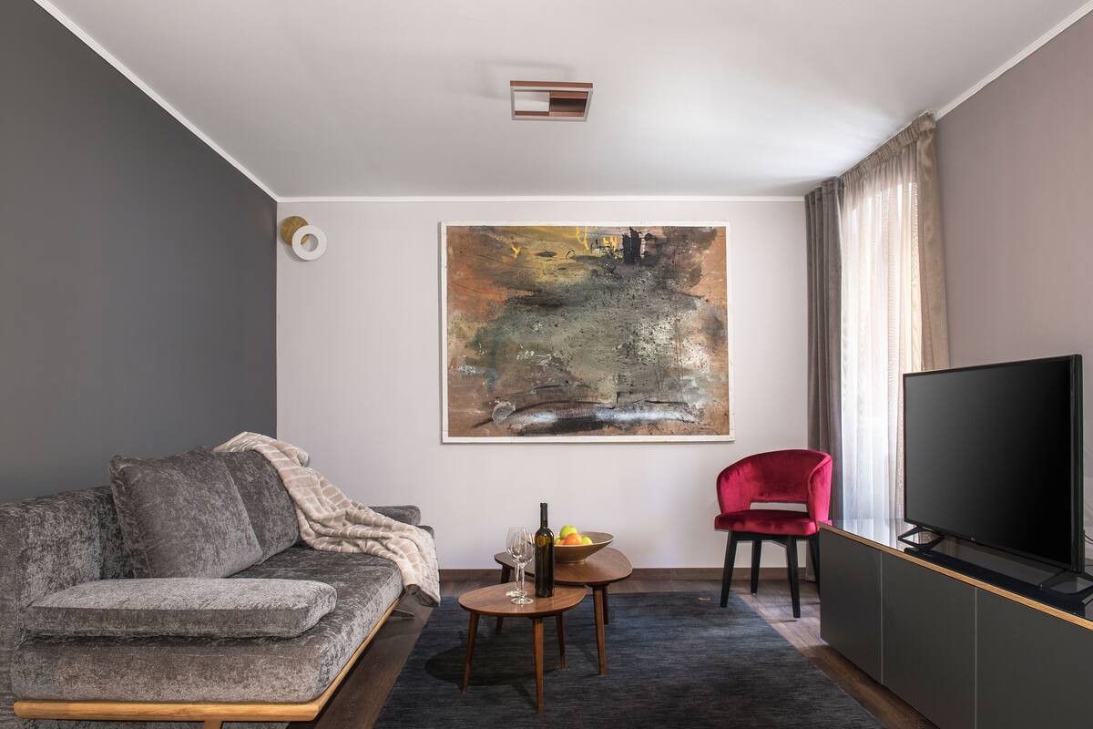 Ein modernes Wohnzimmer mit einem grauen Sofa, einem roten Sessel, einem kleinen Tisch mit einer Obstschale und einer Flasche, sowie einem großen Gemälde an der Wand.