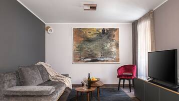 A modern living room with a grey sofa, a red chair, a small table with a bowl of fruit and a bottle, and a large painting on the wall.