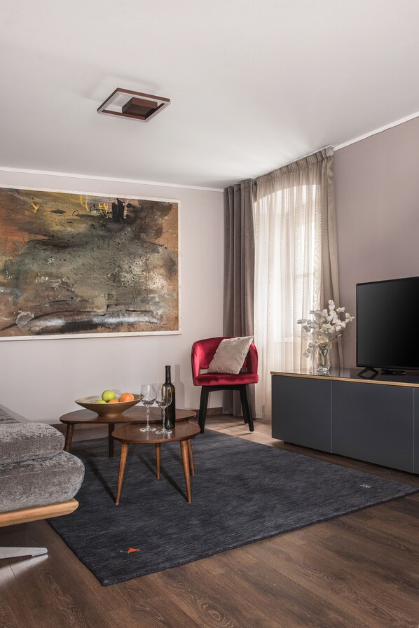 Ein modernes Wohnzimmer mit einem großen abstrakten Gemälde, einem roten Sessel, einem Holztisch mit Früchten und einer Weinflasche sowie einem Flachbildfernseher auf einem dunklen Schrank.