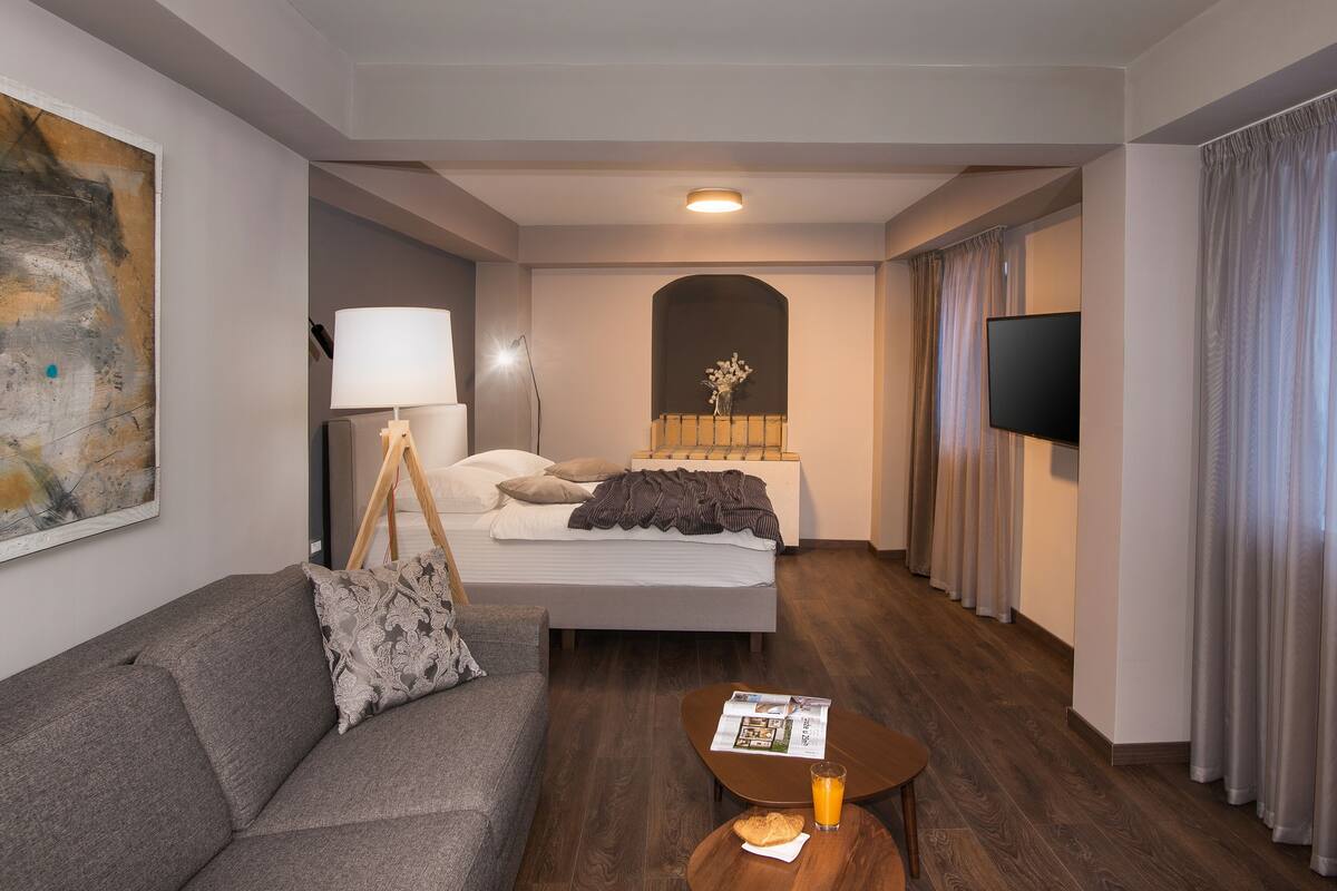 Ein modernes Hotelzimmer mit einem Bett, einem Sofa, einem Couchtisch mit einer Zeitung und einem Glas Orangensaft, einer Lampe und einem Flachbildfernseher.