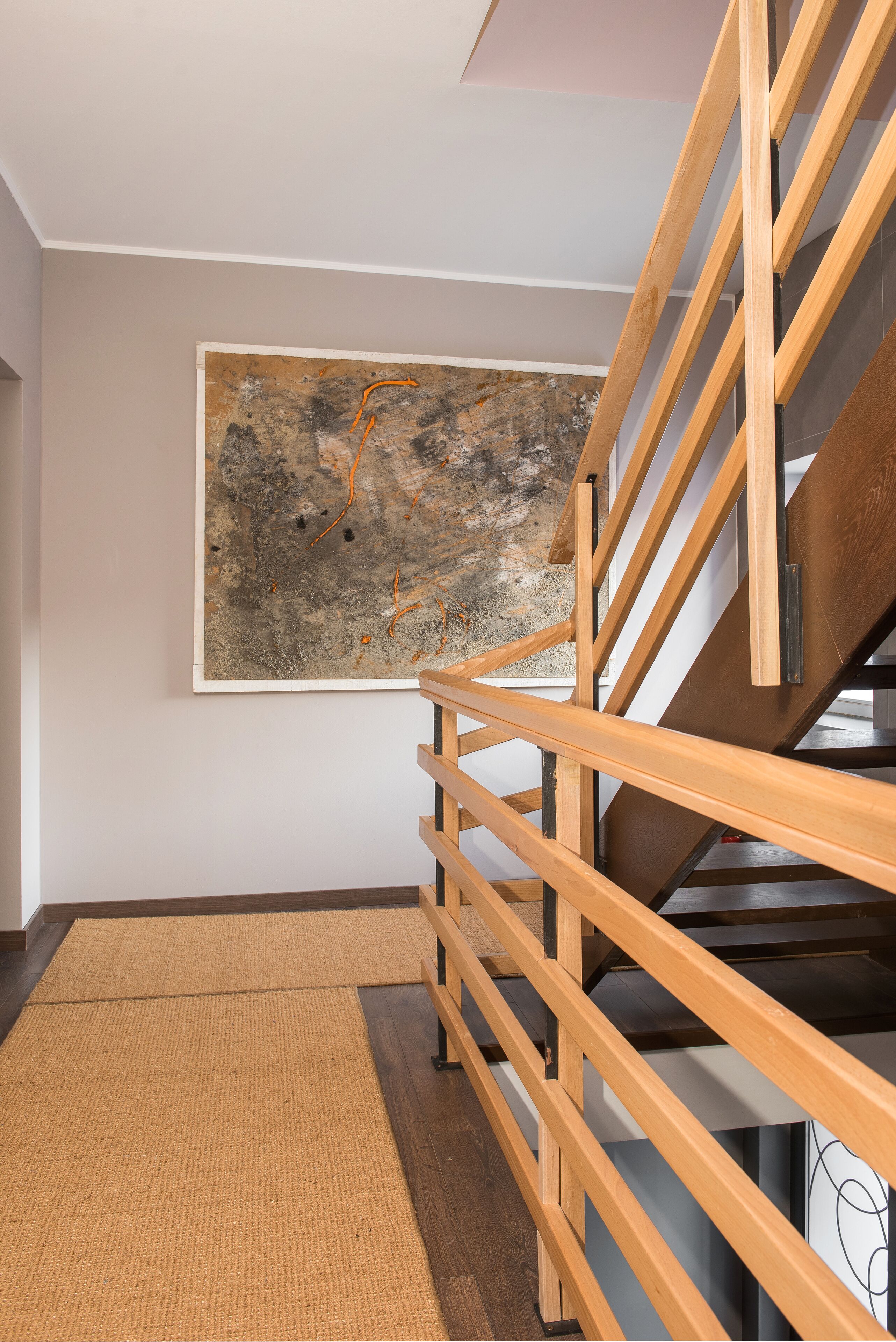 Un interno moderno con una scala in legno, un'ampia pittura astratta appesa al muro e un tappeto dal motivo decorativo sul pavimento.