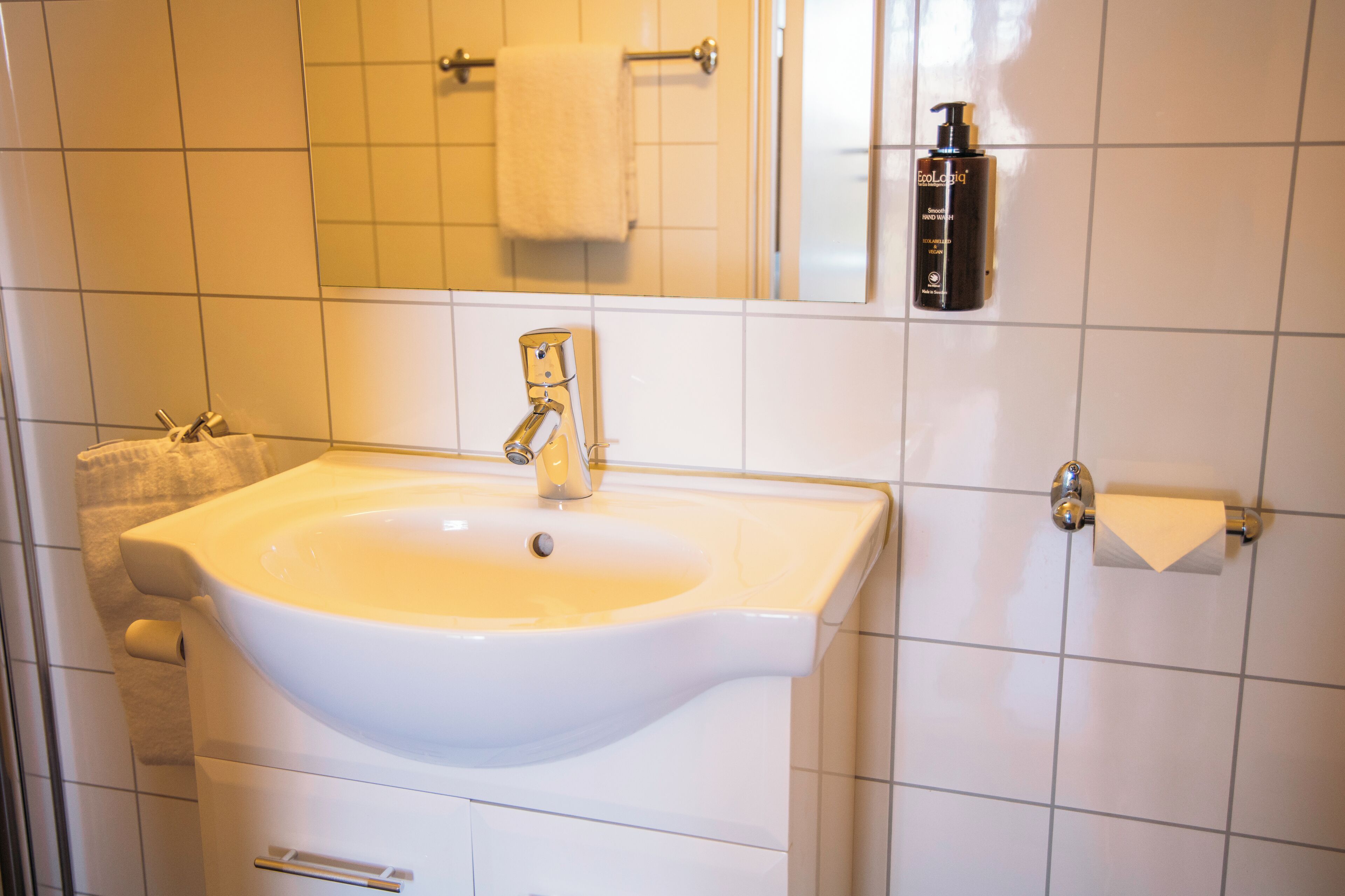 Un baño con lavamanos blanco, toallero, espejo y dispensador de jabón para manos.