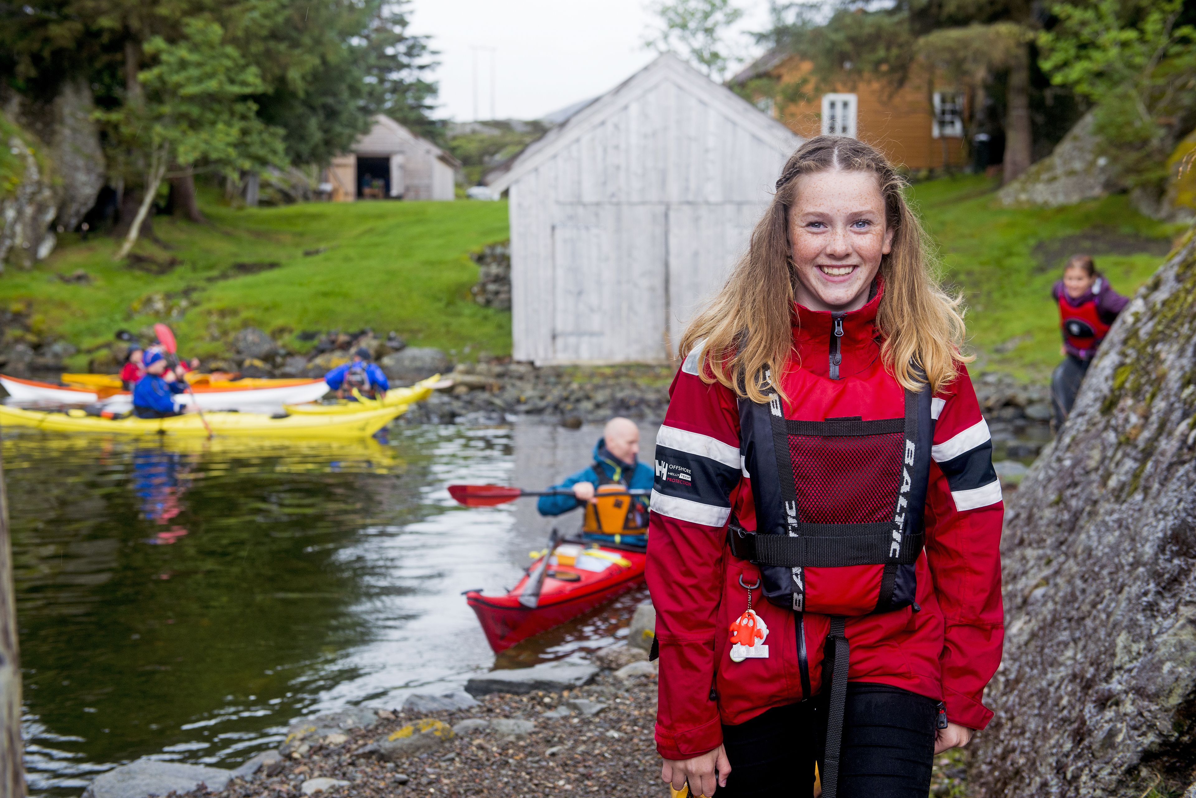 A person in a red jacket with a life jacket standing by a river with kayaks.