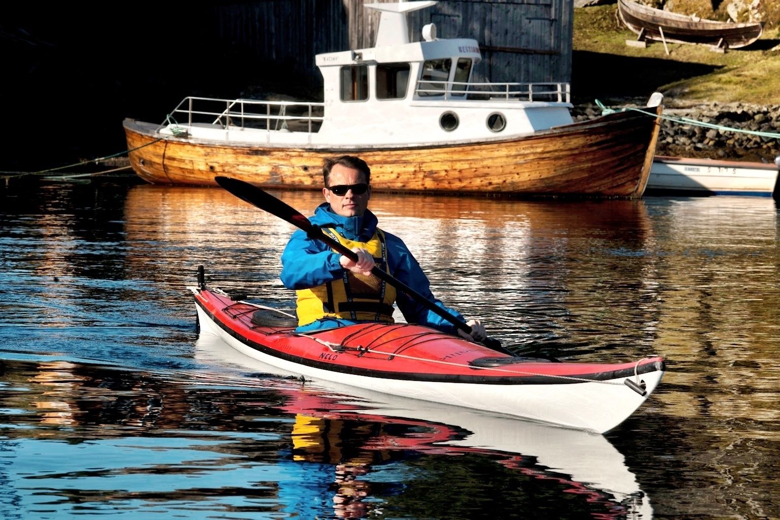 Een persoon in een rode kajak op rustig water, met een houten boot op de achtergrond.