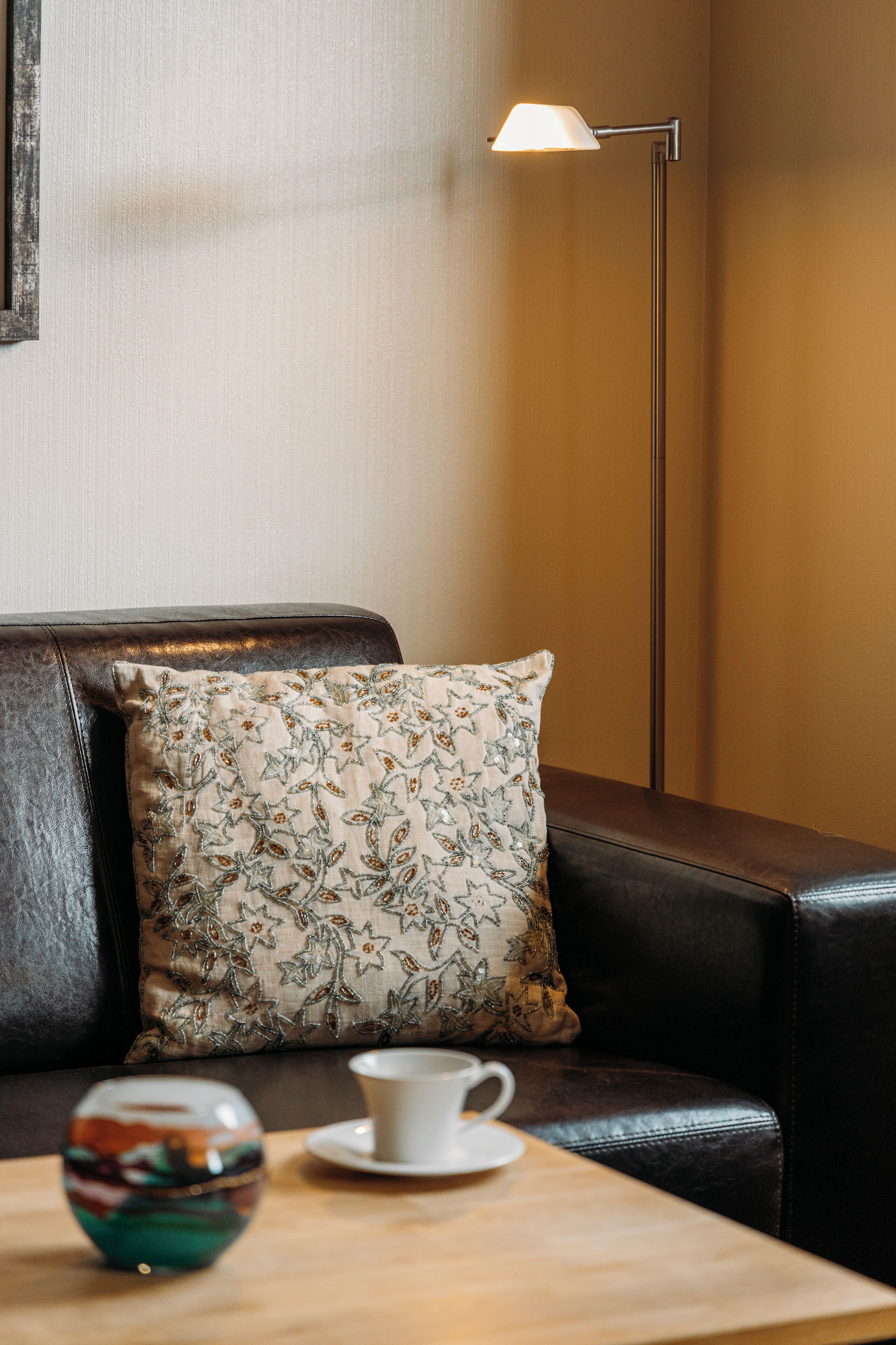 A brown leather sofa with a floral pillow, a white cup and saucer, and a colorful glass bowl on a wooden table.