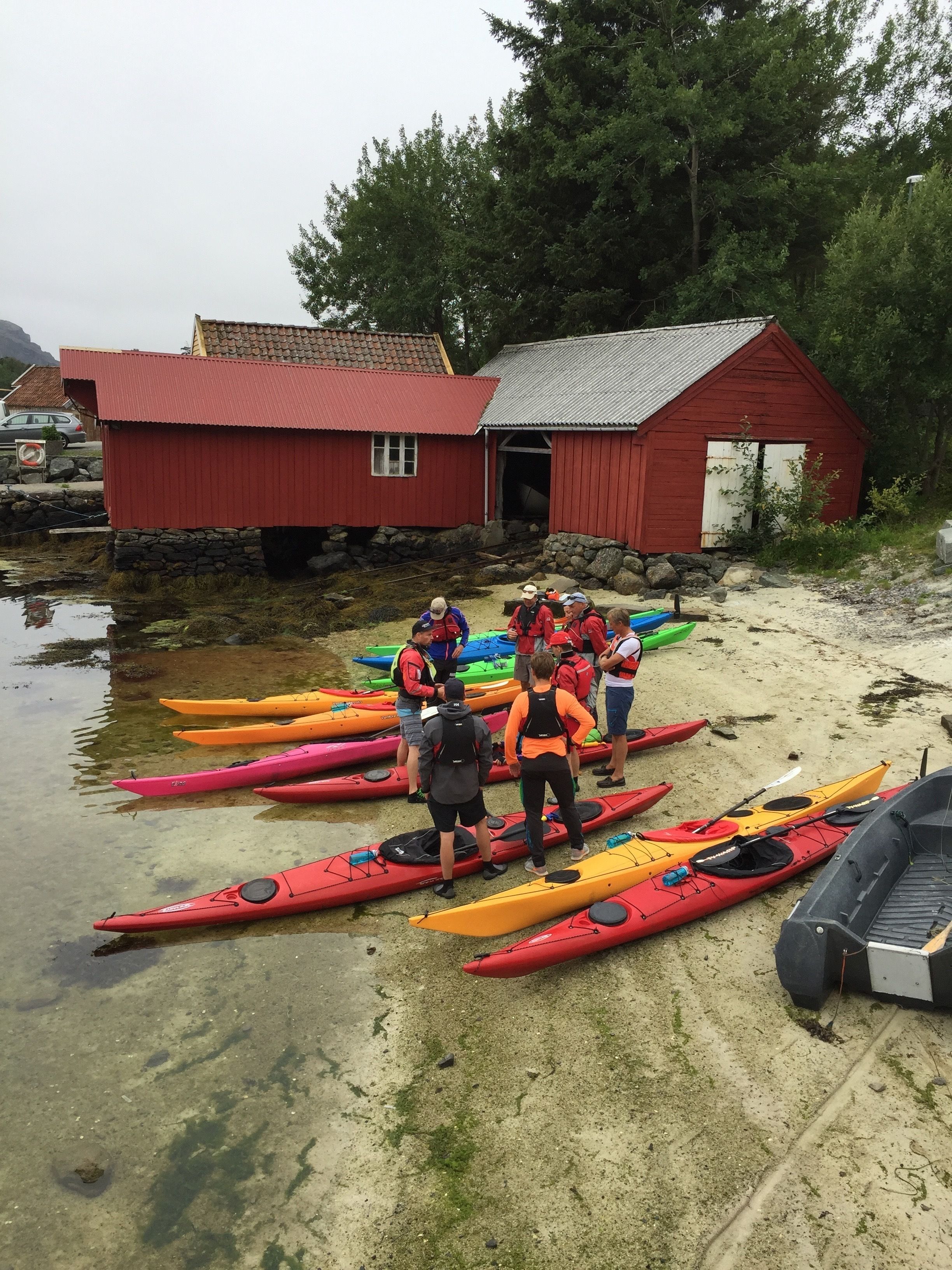 A group of people standing by kayaks near a red building with a stone foundation.