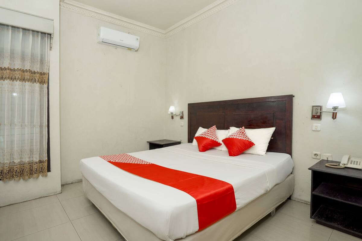 A hotel room with a large bed, a bedside table with a lamp, a phone, and a wall-mounted air conditioner.
