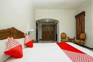 A hotel room with a bed, two red pillows, a wooden headboard, a wardrobe, and two chairs.