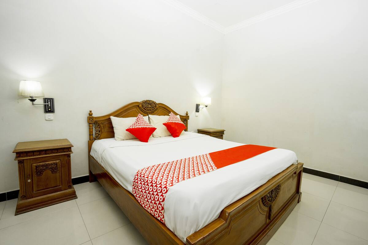 A neatly made bed with a white and red bedspread, two red pillows, and a wooden headboard. There are two bedside tables with lamps on either side of the bed.