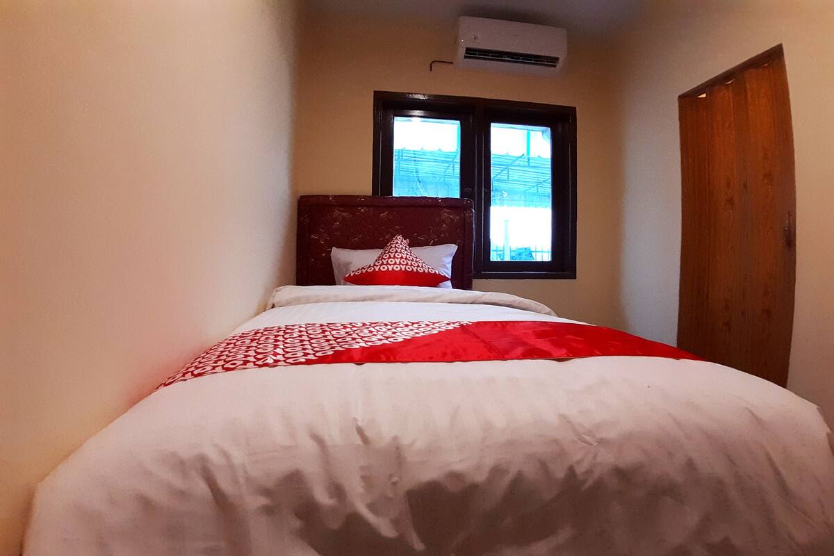 A neatly made bed with a red and white patterned blanket, a wooden headboard, and a window with a view of a building outside.