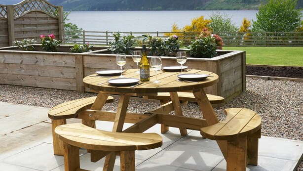 A wooden picnic table with benches, set on a patio with a view of a lake and mountains.