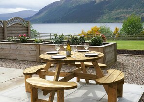 A wooden picnic table with benches, set on a patio with a view of a lake and mountains.