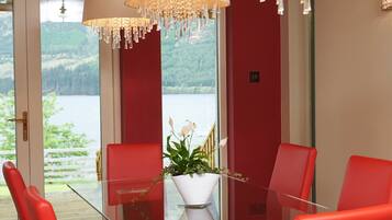 A dining area with a glass table, red chairs, a white vase with a plant, and a view of a lake through large windows.