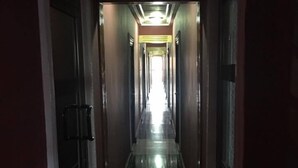 Um corredor comprido e estreito com portas em ambos os lados, dando acesso a quartos. O chão é brilhante e reflete a luz, e a iluminação é forte no final do corredor.