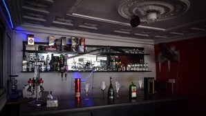 Uma área de bar com prateleiras cheias de garrafas e copos, teto decorativo e um ventilador na parede.
