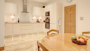 A modern kitchen with white cabinetry, a stainless steel range hood, and a wooden dining table with a fruit bowl. - Luxury Apartments - Rodney Road (Cheltenham)