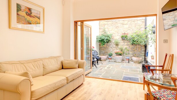 A living room with a beige sofa, a glass table, and chairs, a stone wall, and a door leading to an outdoor area with potted plants. - Luxury Apartments - Rodney Road (Cheltenham)