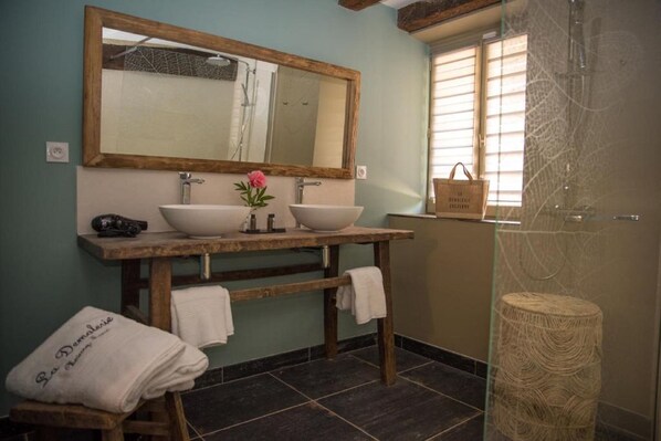Un baño con dos lavamanos, un espejo grande y una ducha. Hay toallas en un banco y una pequeña cesta en la pared.