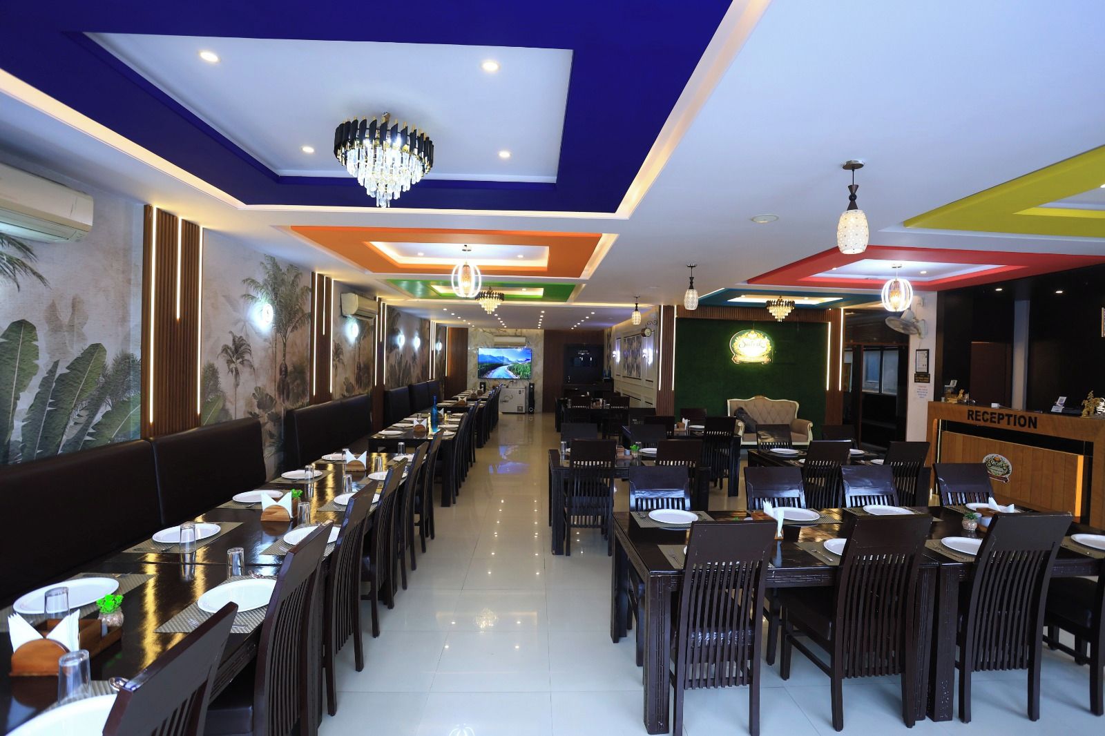 Un ristorante con diversi tavoli e sedie, una reception e uno schermo televisivo sullo sfondo.