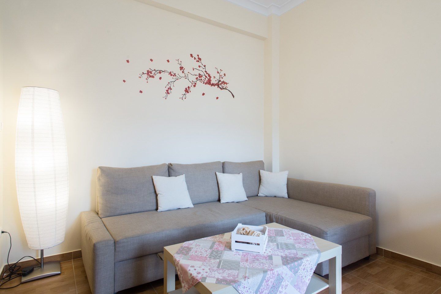 A modern living room with a grey sofa, white cushions, a small table with a floral tablecloth, and a wall decoration of red branches.