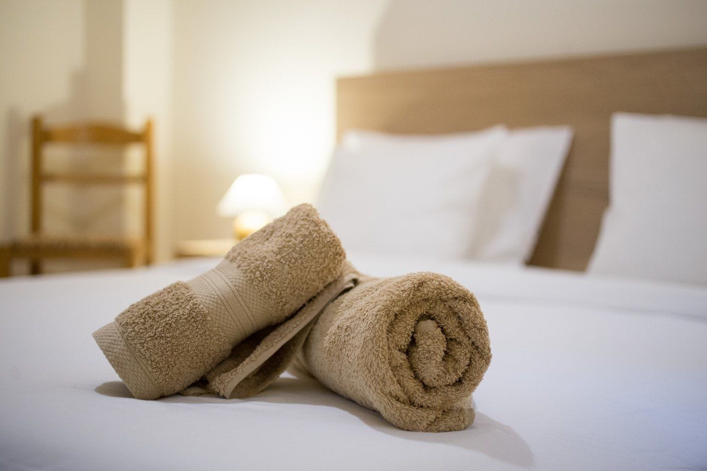 Een net opgemaakt bed met opgerolde handdoeken en een houten stoel in een hotelkamer.