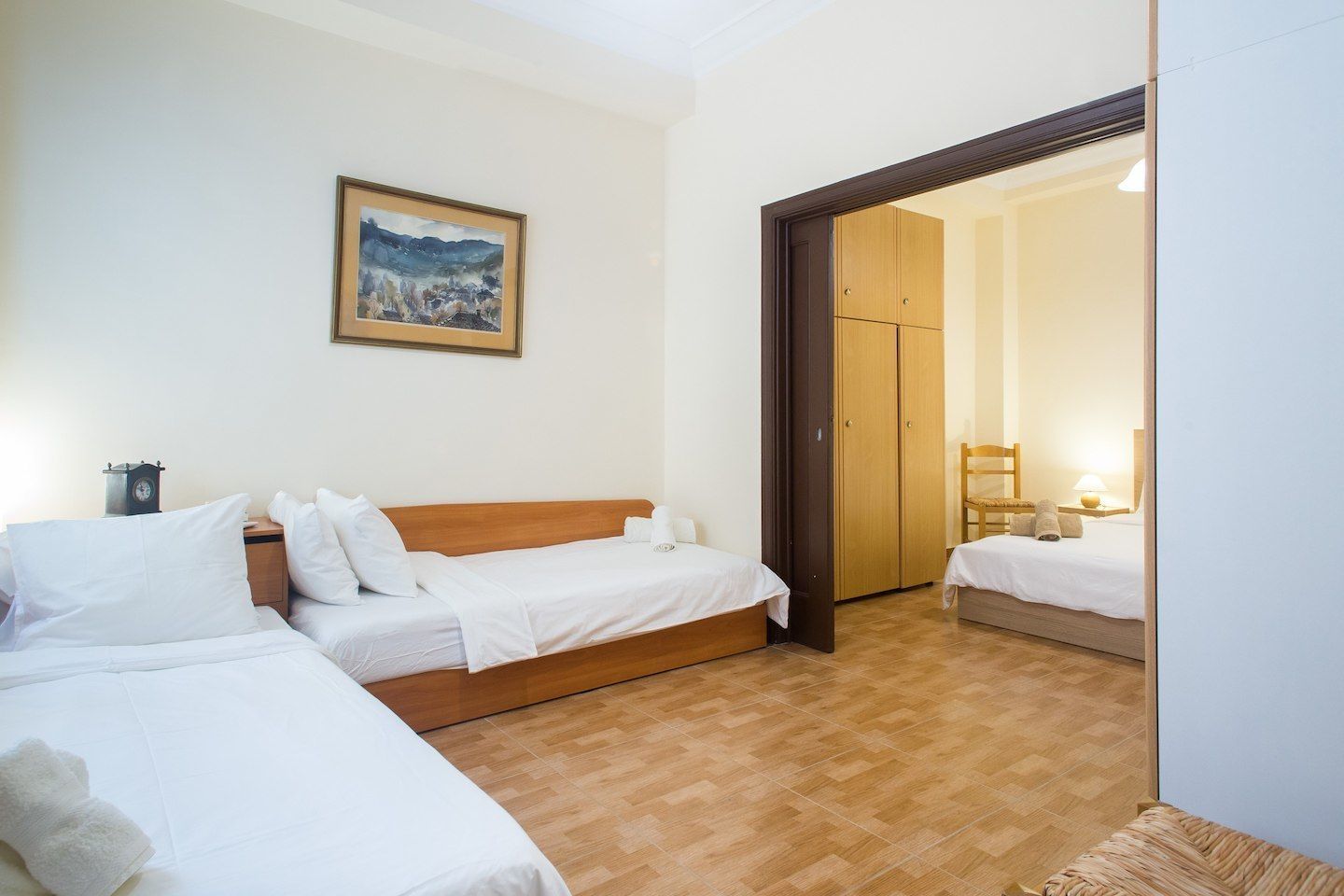 Habitación de hotel con dos camas individuales, cabecera de madera, cuadro enmarco con un paisaje, armario de madera y una mesita con lámpara.