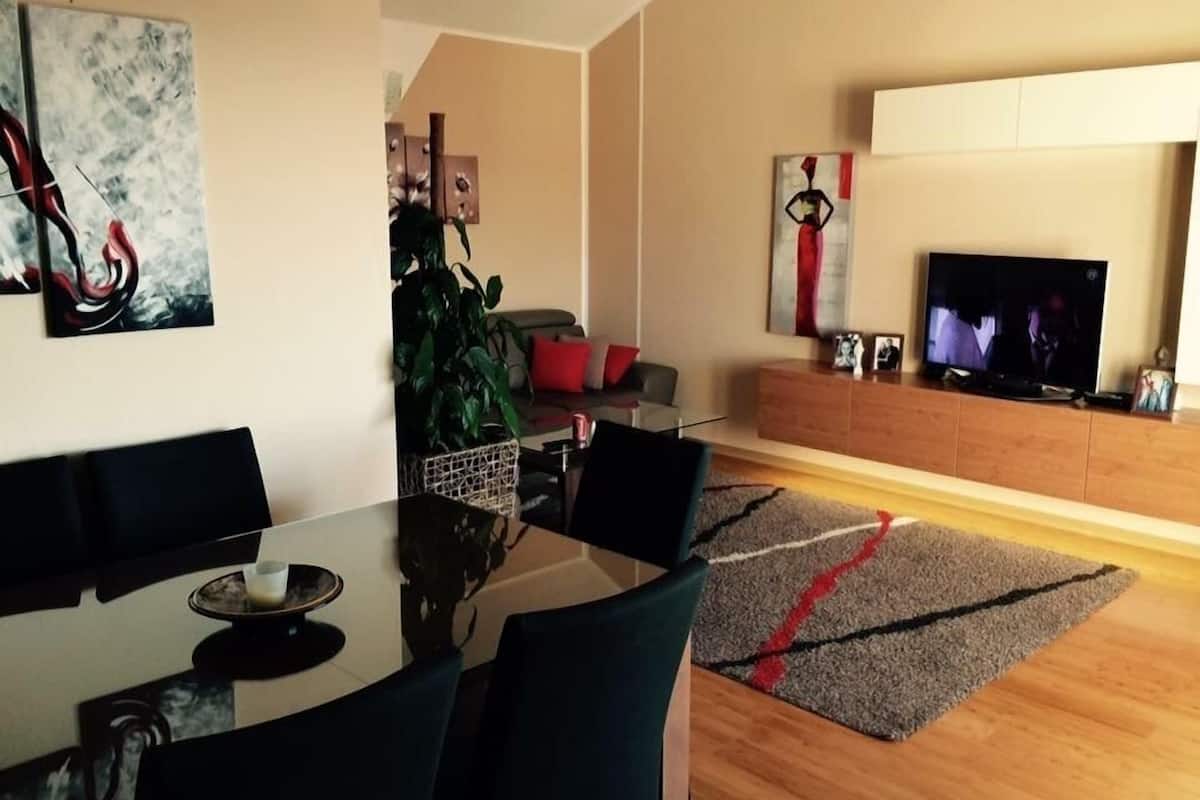 A modern living room with a glass dining table, black chairs, a sofa, a TV, and artwork on the walls.
