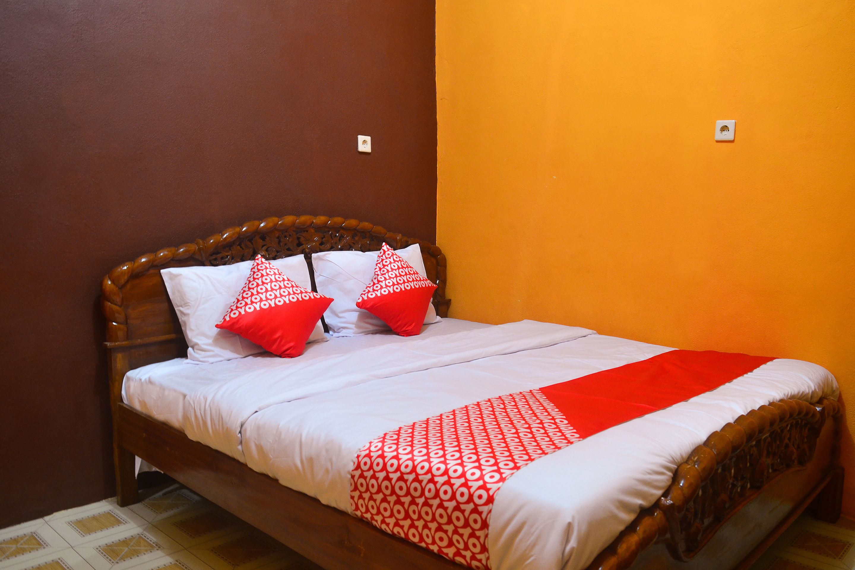 Uma cama com cabeceira de madeira, roupa de cama branca e almofadas vermelhas estampadas.