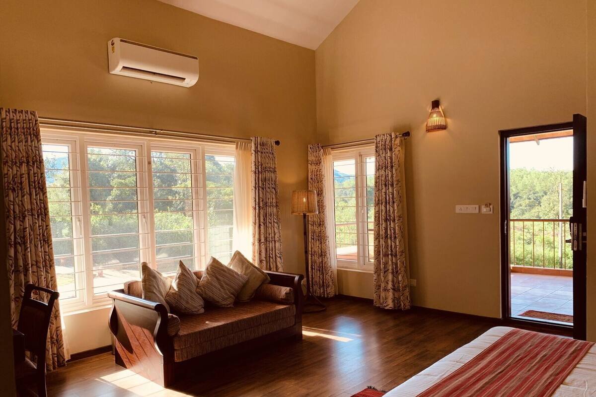 A room with a wooden floor, a sofa with cushions, a bed with a striped blanket, and large windows with curtains.