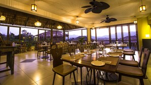 A spacious dining area with multiple tables and chairs, large windows offering a view of the mountains, and ceiling fans.