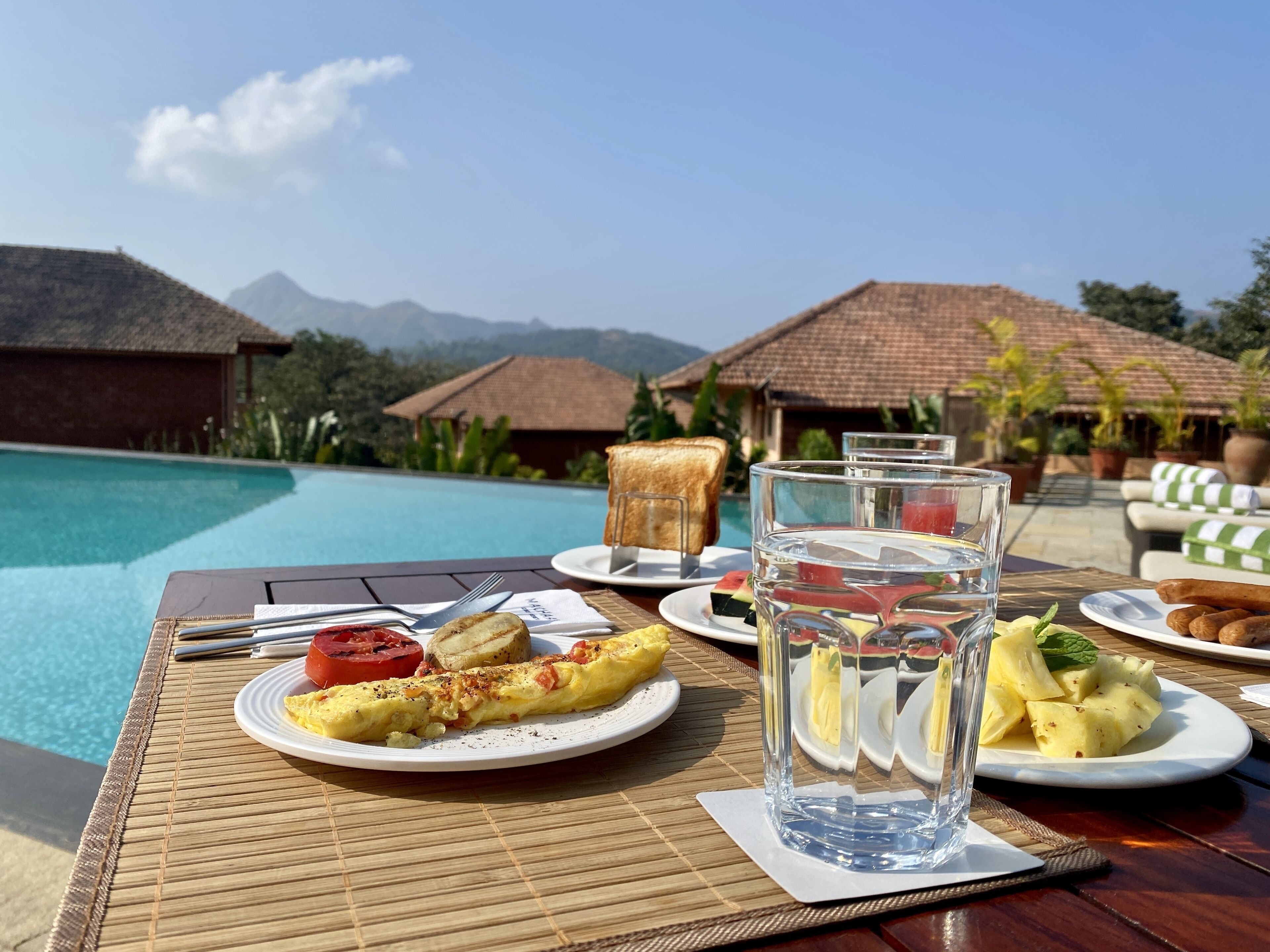 Ein Frühstückstisch mit Blick auf einen Pool, mit Speisen, Gläsern Wasser und tropischer Landschaft im Hintergrund.