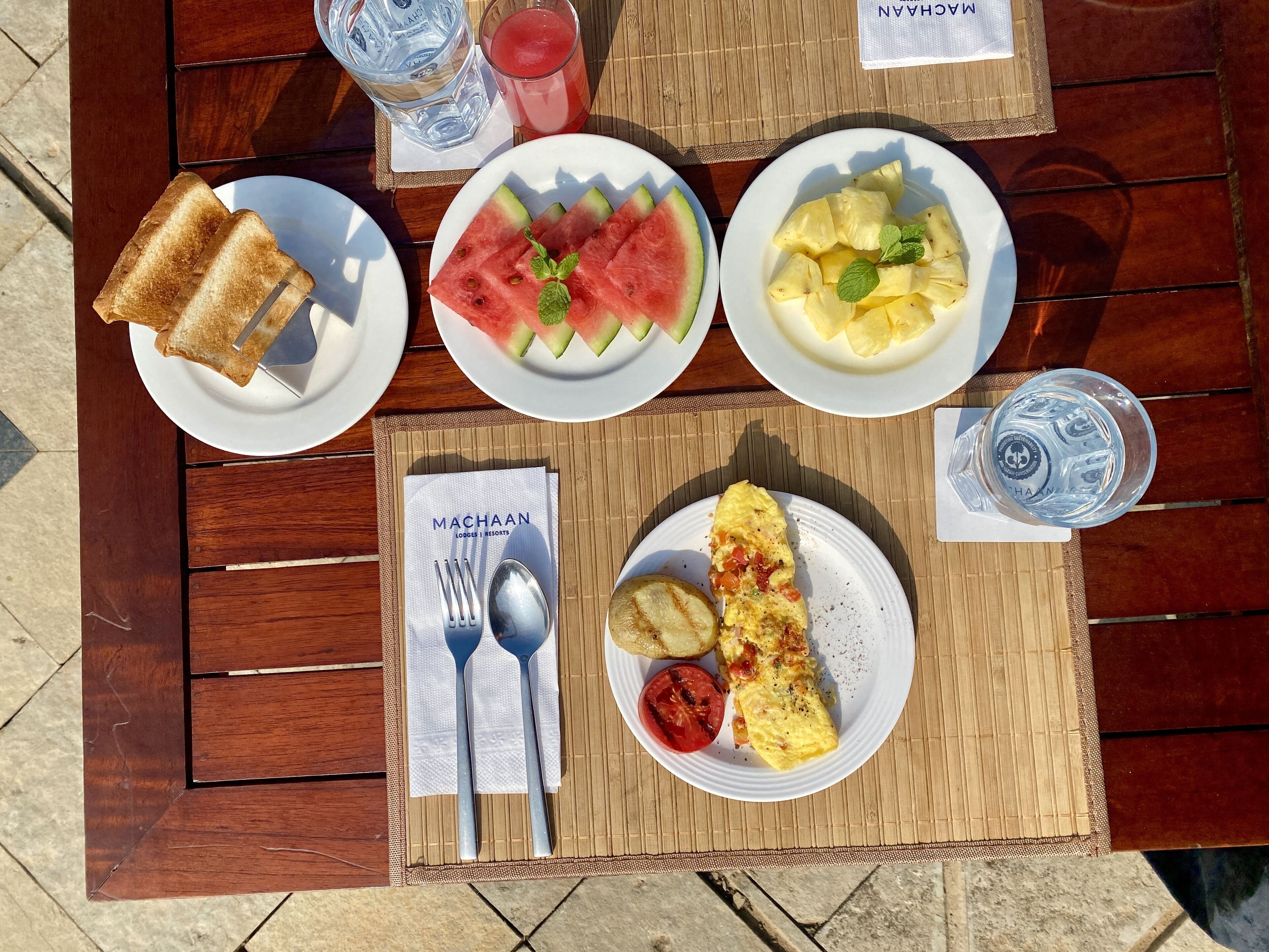 Una mesa dispuesta con alimentos para el desayuno, como tostadas, sandía, piña, una tortilla y una bebida.