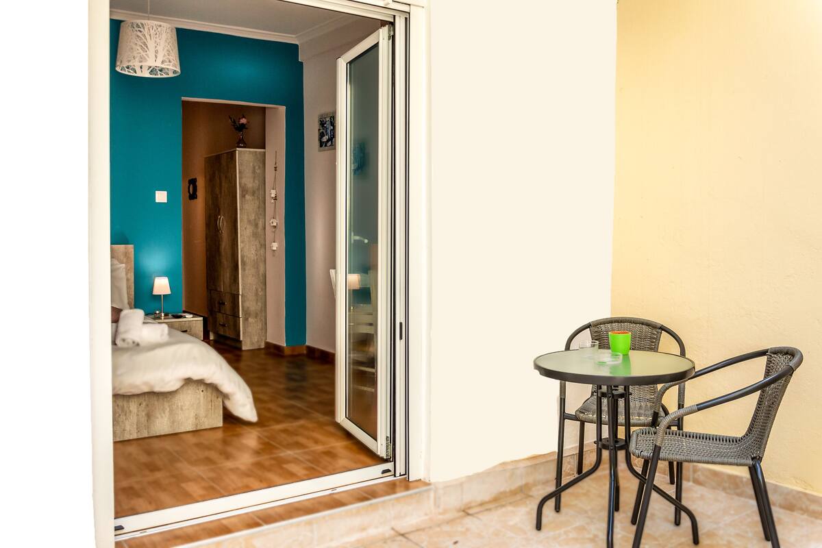 Una habitación con cama, una mesa con sillas y un ventanal corredero que da a otra habitación.